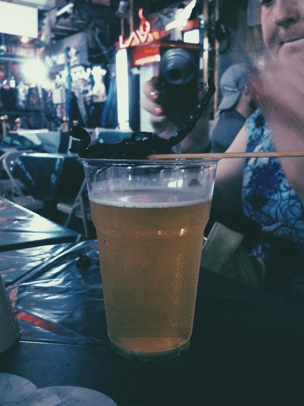 Colored Photograph of a Scorpion Skewer and a Cup of Beer at a Bar on Khao San Road.